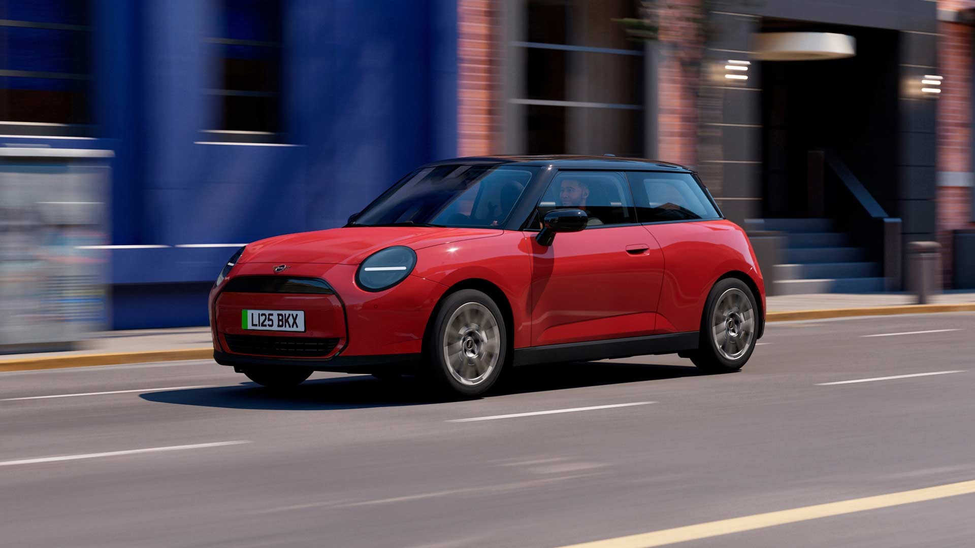 MINI Cooper electric hatchback driving on a city street with blue and brick buildings in the background, featuring black roof, circular LED headlights, and sleek modern design.